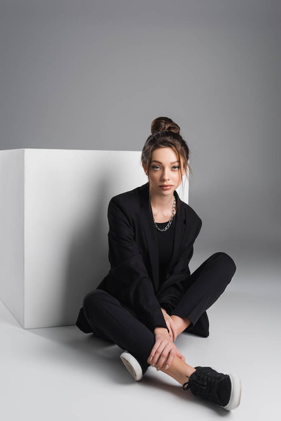 stylish woman in black suit and sneakers sitting with crossed legs near white cube on grey background - Foto, afbeelding