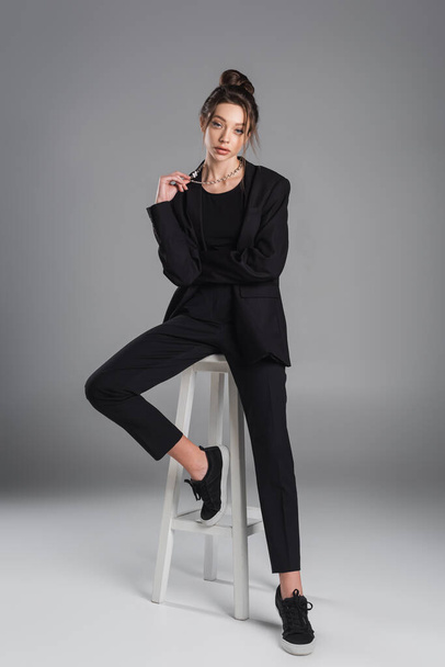 full length of woman in black clothing touching silver necklace while posing on white stool on grey background - Photo, Image