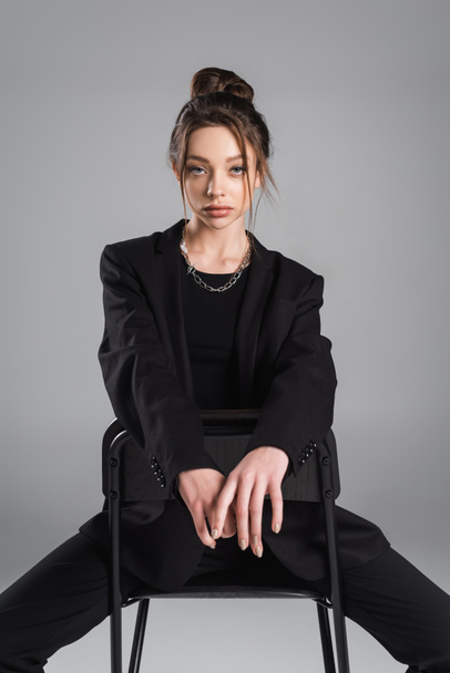 brunette woman in black clothes sitting on chair and looking at camera isolated on grey - Photo, Image
