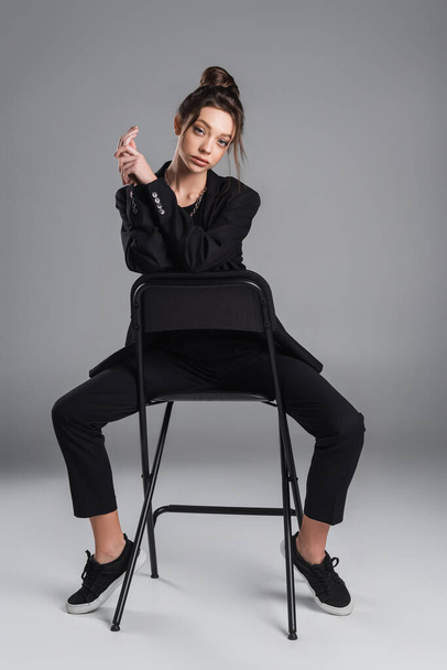 full length of pretty woman in black suit and sneakers posing on chair on grey background - Foto, Imagem