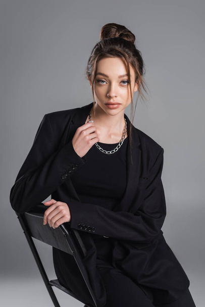 fashionable woman in black suit and silver necklace looking at camera while sitting isolated on grey - Valokuva, kuva