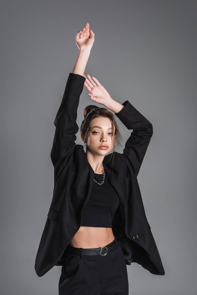 young woman in black crop top and blazer posing with raised hands isolated on grey - 写真・画像
