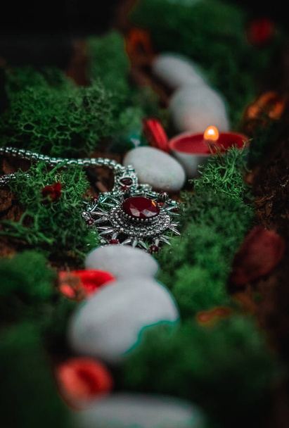 A ritual amulet with a red gem, green moss, a candle, dried flowers and gray stones in the bark of a tree lies diagonally on a black background, close-up side view. Esoteric concept, dark style. - Foto, immagini