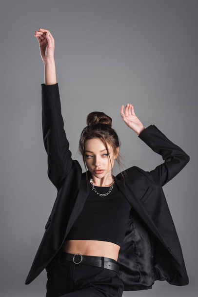 trendy woman in crop top and black jacket standing with arms up isolated on grey - Foto, afbeelding