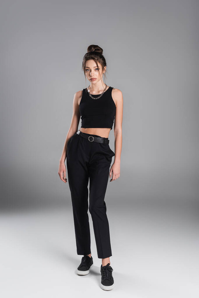 full length of young woman in black trousers and sneakers standing on grey background - Foto, Imagem