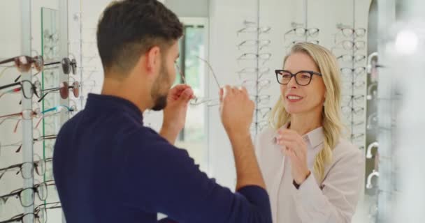 Female optician or optometrist assisting a customer to choose glasses on the shelf in her shop. Friendly mature shop owner helping a man to choose and try on spectacles in her optical store. - Séquence, vidéo