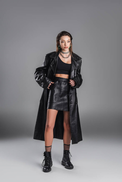 full length of young woman in black leather coat standing with hand in pocket of skirt on grey background - Foto, afbeelding
