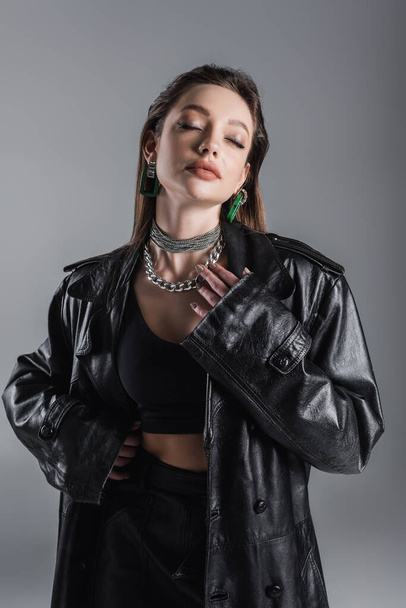 sensual woman in leather clothes and silver necklaces posing with closed eyes isolated on grey - Photo, Image