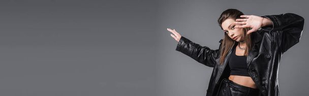 pretty woman in black crop top and leather coat gesturing and looking at camera isolated on grey, banner - Foto, imagen