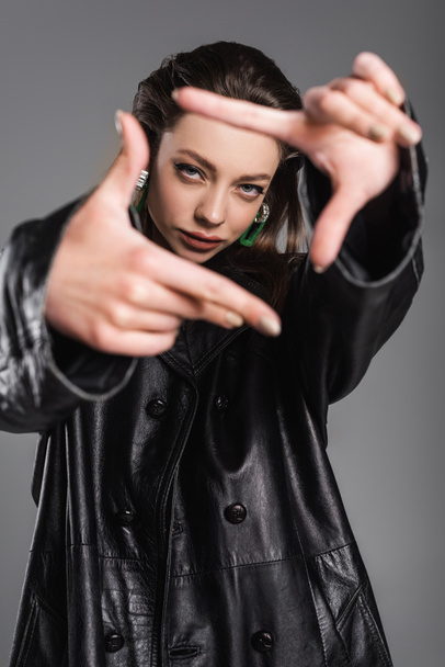 trendy woman in black leather coat looking at camera and gesturing on blurred foreground isolated on grey - Foto, immagini