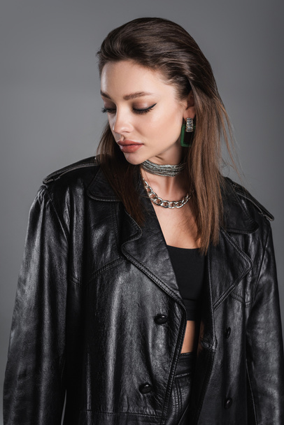 young woman with makeup and silver necklaces posing in black leather coat isolated on grey - Φωτογραφία, εικόνα