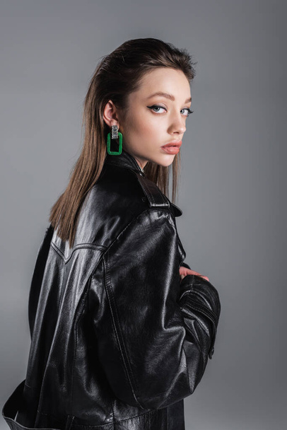 brunette woman with makeup standing in black leather coat and looking at camera isolated on grey - Фото, зображення