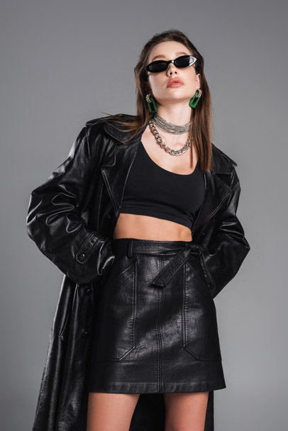 young woman in dark sunglasses and black leather clothing posing with hands in pockets isolated on grey - Фото, изображение