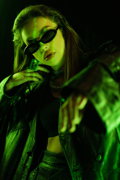 trendy woman in dark sunglasses and leather coat gesturing in green light isolated on black - Photo, image