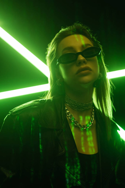 low angle view of woman in dark sunglasses on black background with green neon light - Photo, Image