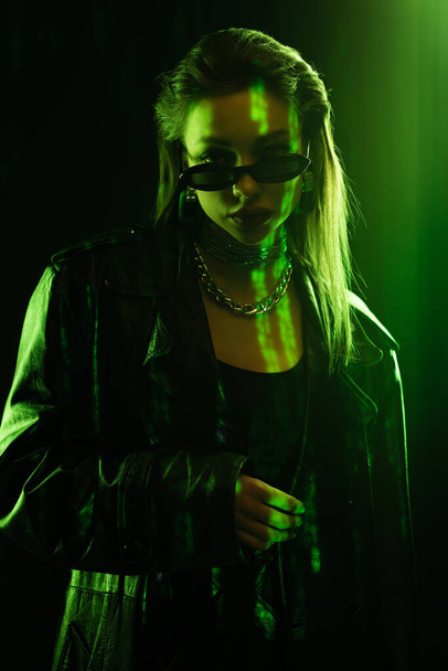 fashionable woman in sunglasses and leather coat looking at camera on black background with green light - Foto, Imagen