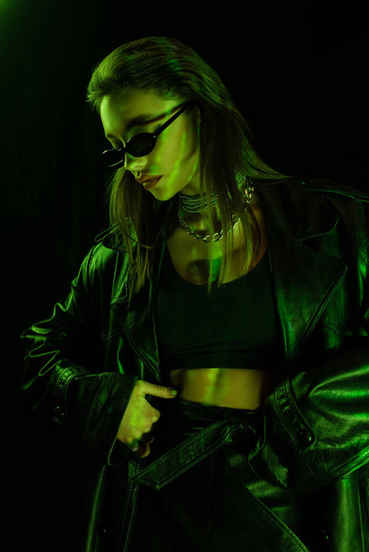 trendy woman in sunglasses and crop top standing with hand on hip in green light isolated on black - Fotografie, Obrázek