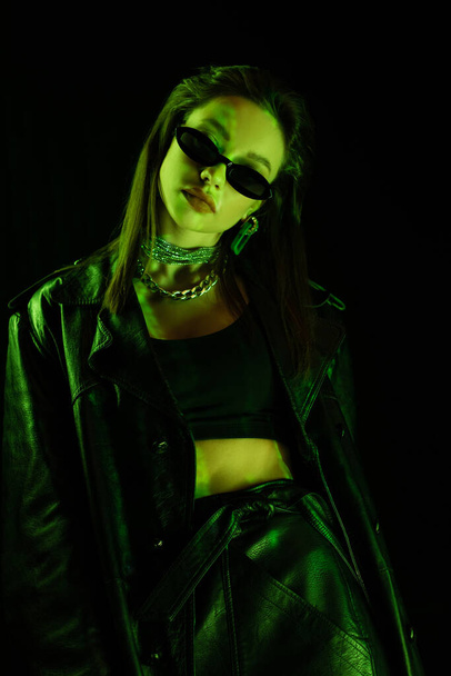 модна жінка в шкіряному одязі та сонцезахисних окулярах у зеленому неоновому світлі ізольована на чорному
 - Фото, зображення