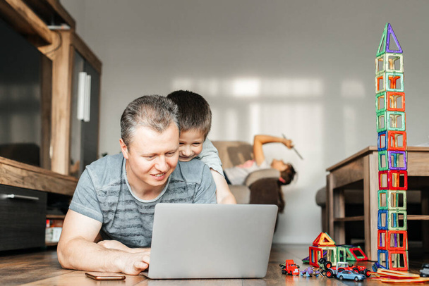 Papa werkt thuis op een laptop en speelt tegelijkertijd met zijn zoon. Het kind knuffelt vader en bemoeit zich met het werk. Kinderen en thuiswerk op afstand. - Foto, afbeelding