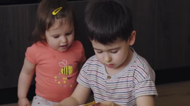 Caucasian brother and baby sister looking and smiling expression while reading a magazine indoor home sitting on the floor. Smiling happy child. Family care. - Filmagem, Vídeo
