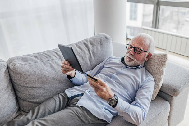 Thoughtful senior executive analyzing digital tablet. Freelancer making online payment through credit card. He is lying on sofa at home office. - Foto, immagini