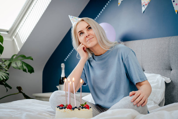Thoughtful smiling woman in birthday party cap with festive cake with candles on the bed in decorated bedroom. Make a wish process. Morning surprise. Happy birthday concept. Selective focus. - Zdjęcie, obraz
