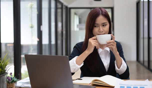 Hermosa joven empresaria asiática sonriendo sosteniendo una taza de café y portátil trabajando en la oficina - Foto, imagen
