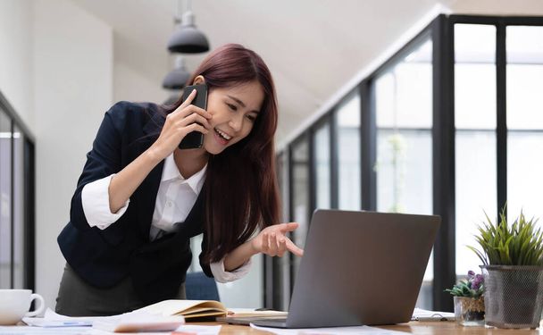 Hermosa atractiva joven mujer de negocios asiática hablando por teléfono con su colega y comprobando la información en la pantalla del ordenador portátil - Foto, imagen