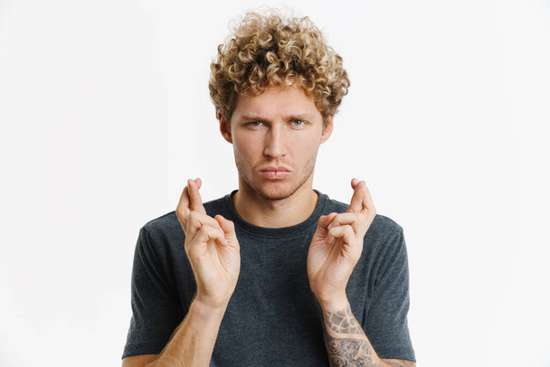 Young blonde man holding fingers crossed for good luck isolated over white wall - Foto, afbeelding