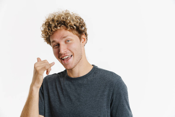 Young blonde man with curly hair smiling and making call gesture isolated over white wall - Photo, image