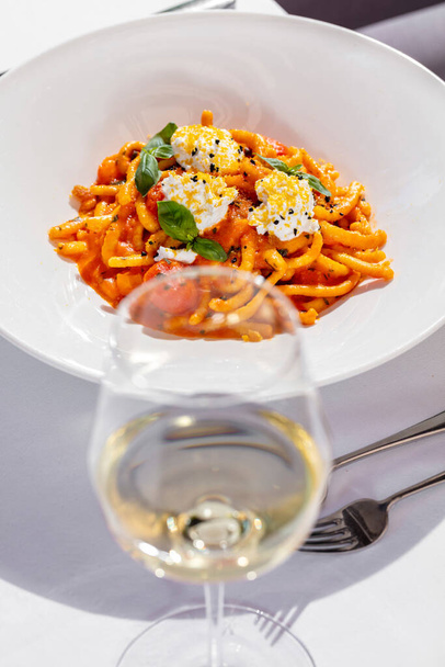 Italian pasta Margherita with tomato sauce, basil and mozzarella. Pasta lies in a light, ceramic plate. Nearby is a glass of white wine and cutlery. Food and wine on a table with a white tablecloth. - Fotografie, Obrázek