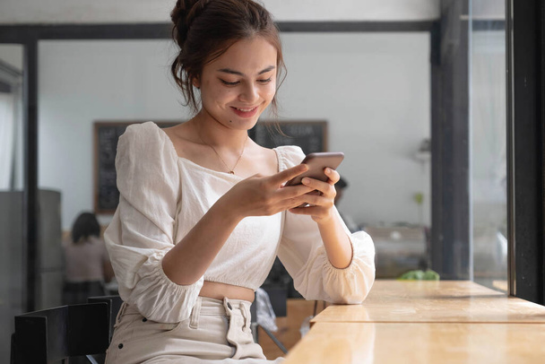 Азиатская девушка использует смартфон в кафе. Улыбающаяся женщина, пользующаяся смартфоном дома, отправляющая сообщения или просматривающая социальные сети во время отдыха - Фото, изображение