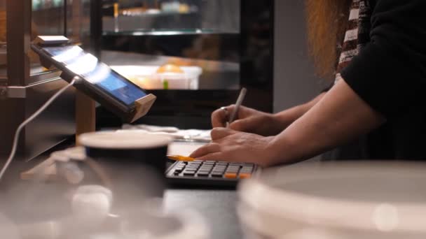 Male clerk makes calculations on a calculator and writes, behind the counter in a small shop. Man makes notes on documents with a pen - Séquence, vidéo