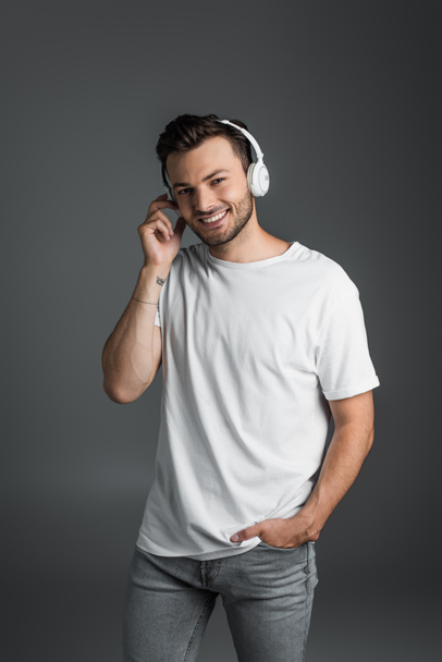 Smiling man in t-shirt and jeans using headphones isolated on grey  - Photo, Image