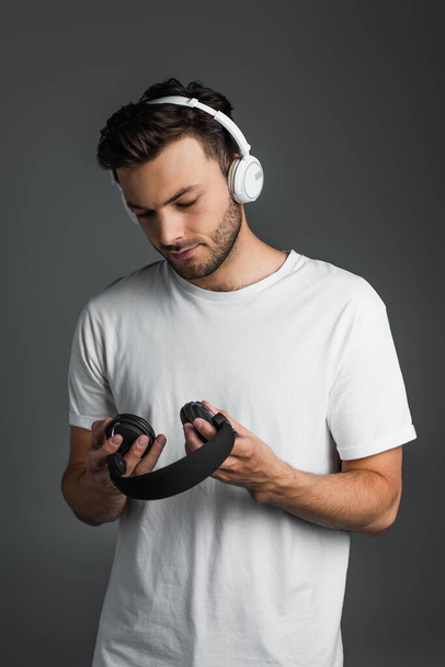 Brunette man in t-shirt looking at wireless headphones isolated on grey  - Zdjęcie, obraz