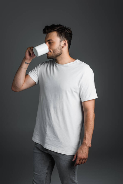 Young man in jeans and t-shirt drinking coffee isolated on grey  - Foto, Imagen