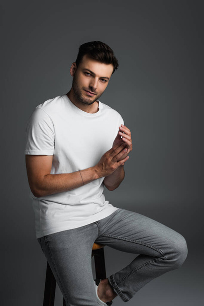 Brunette man in jeans and t-shirt posing while sitting on chair isolated on grey  - Foto, Imagem