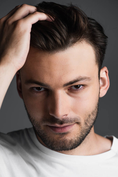 Portrait of young man touching hair and looking at camera isolated on grey  - Foto, Imagen