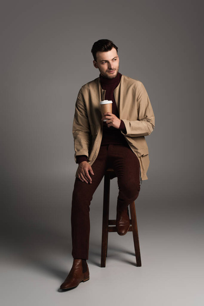 Full length of stylish man in autumn outfit holding paper cup while sitting on chair on grey background - Foto, Imagem