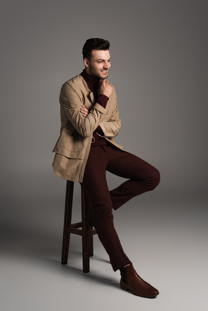 Full length of smiling man in autumn outfit sitting on chair on grey background - Foto, imagen