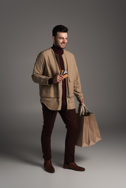 Full length of smiling man in autumn outfit holding shopping bags and credit cards on grey background - Photo, image