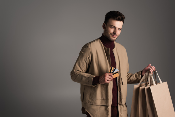 Man in brown jacket holding shopping bags and credit cards isolated on grey  - Fotoğraf, Görsel