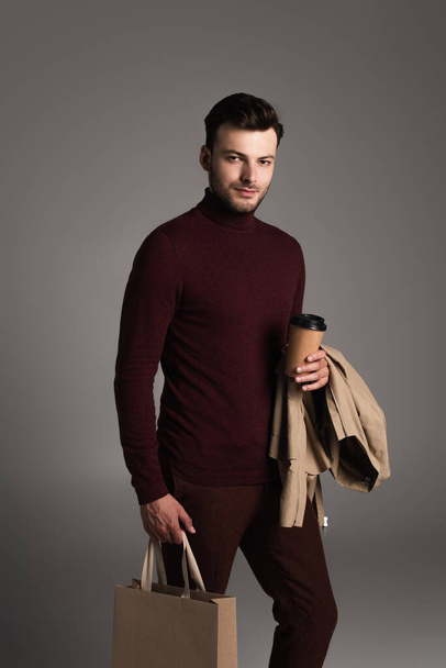 Stylish brunette man with jacket holding coffee to go and shopping bag isolated on grey  - Photo, Image