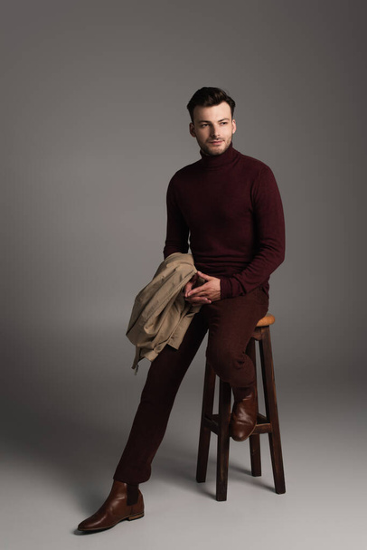 Full length of young barded man in turtleneck holding jacket while sitting on chair on grey background - 写真・画像