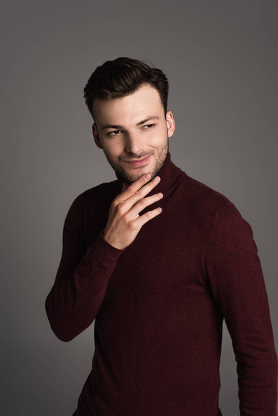 Smiling brunette man in turtleneck touching chin isolated on grey  - Fotografie, Obrázek