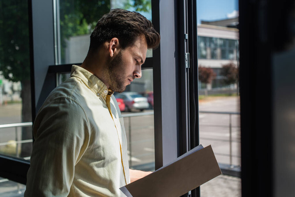 Side view of businessman looking at paper folder near window in office  - 写真・画像