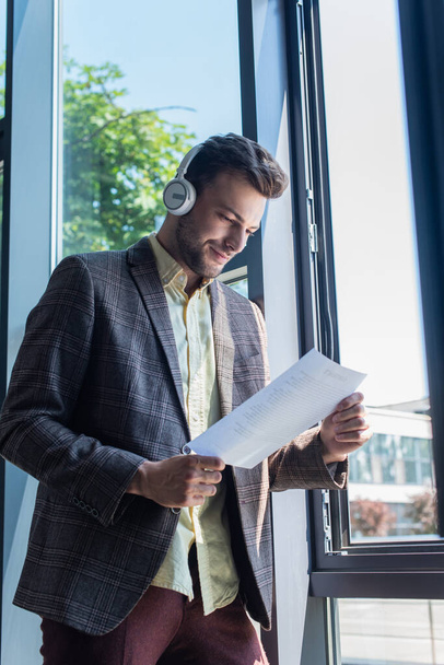 Young businessman in headphones looking at paper near window in office  - Foto, afbeelding