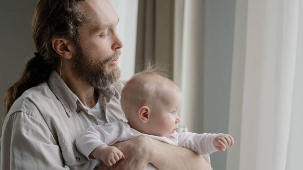 Close-up caucasian father bearded daddy holding baby daughter son infant newborn looking out window with curtains dad blowing to short blonde hair of kid child playing at home. Fatherhood protection - Photo, Image