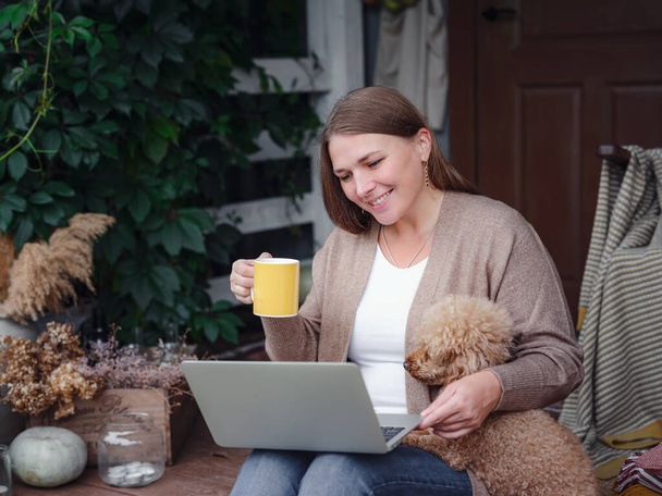 middelbare leeftijd Kaukasische vrouw in bruin vest zit op het terras van het huis met haar beige poedel met laptop werken buiten in de tuin, thuis kantoor, Onderwijs, moderne levensstijl en vrije tijd concept. - Foto, afbeelding
