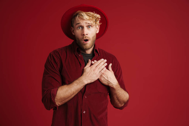 White shocked man wearing hat holding hands on his heart isolated over red wall - Foto, immagini